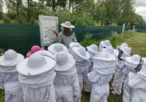 Dzieci ubrane w kombinezony pszczelarskie słuchają pana pszczelarza opowiadającego o cyklu rozowju pszczół.