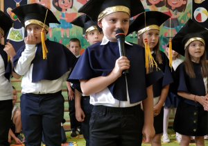 Przedszkolaki podczas występu zakończenia roku szkolnego. Chłopic mówi wiersz do mikrofonu.