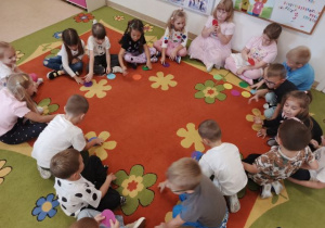 Przedszkolaki siedzą w kole na dywanie i uczestniczą w zabawie z kolorowymi kropkami.