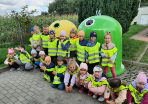 Przedszkolaki pozują do zdjęcia, w tle widać pojeminki do segregacji odpadów.