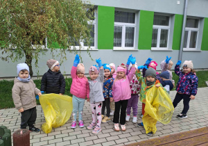 Dzieci z grupy 3-4 latków stoją przed przedszkolem z rękawiczkami i workami na śmieci przygotowują się do akcji