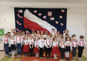 Przedszkolaki śpiewają hymn na tle biało-czerwonej flagi.