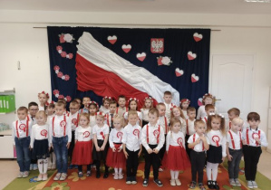 Przedszkolaki śpiewają hymn na tle biało-czerwonej flagi.