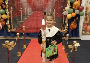 Dziewczynka trzyma dyplom i "magiczny ołówek", pozuje do zdjęcia na czerownym dywanie.