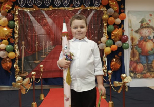 Chłopiec trzyma dyplom i "magiczny ołówek", pozuje do zdjęcia na czerownym dywanie.