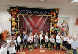 Grupa młodsza stoi w półkolu na tle dekoracji i mówi wierszyk.