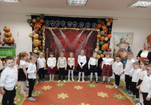 Grupa starsza stoi w półkolu na tle dekoracji i śpiewa piosenkę.