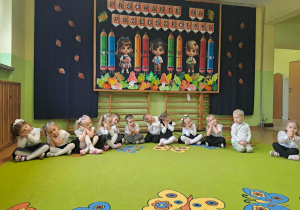 Przedszkolaki siedzą w półkolu na dywanie i pokazują określone gesty do piosenki