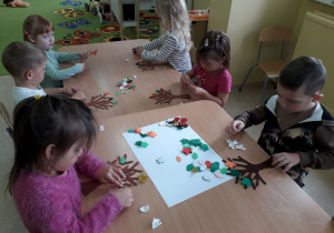 Dzieci przy stoliku ozdabiają drzewo jesiennymi , kolorowymi liśćmi.