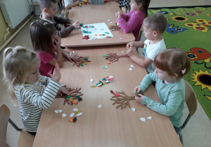 Dzieci przy stoliku ozdabiają drzewo jesiennymi , kolorowymi liśćmi.