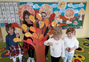 Przedszkolaki ozdabiają duże, papierowe, jesienne drzewo wykonanymi sylwetami żołędzi.