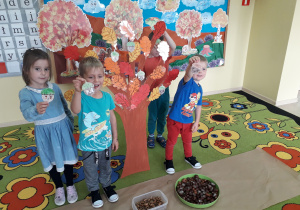 : Przedszkolaki ozdabiają duże, papierowe, jesienne drzewo wykonanymi sylwetami żołędzi.