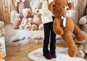 Dziewczynka pozuje do zdjęcia trzymając wielkiego misia i dyplom.