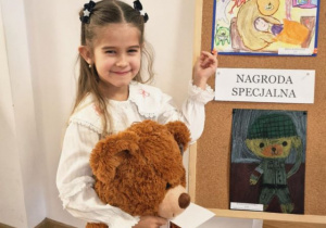 Dziewczynka trzymając wielkiego misia prezentuje swoją pracę konkursową.