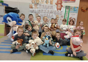 Dzieci siedzą ze swoimi misiami. W tle dekoracja z napisem Dzień Pluszowego Misia.