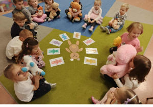 Dzieci siedzą w kole i trzymają swoje misie. Przed nimi karty obrazkowe.