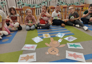 Dzieci siedzą w kole i trzymają swoje misie. Przed nimi karty obrazkowe.