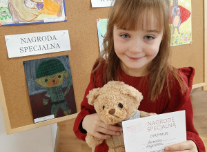Dziewczynka pozuje do zdjęcia z misiem i dyplomem. W tle praca konkursowa.