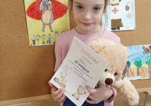 Dziewczynka pozuje do zdjęcia z misiem i dyplomem. W tle praca konkursowa.