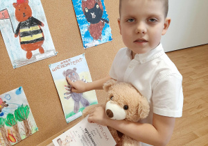 Chłopiec pozuje do zdjęcia z misiem i dyplomem. W tle praca konkursowa.