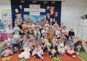 Dzieci trzymają swoje misie i pozują do zdjęcia. W tle tematyczna dekoracja z misiami.