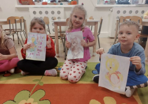 Dwie dziewczynki i chłopic siedzą na dywanie i prezentują narysowane misie.