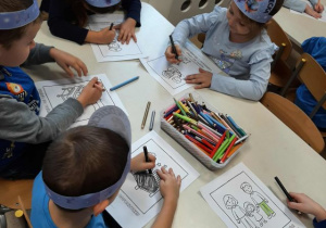 Przedszkolaki siedzą przy stoliku i kolorują obrazki.