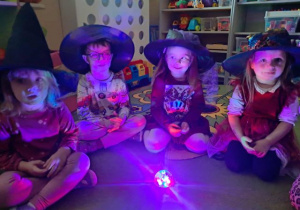 Dzieci w kapeluszach siedzą na dywanie przy kolorowej lampce.