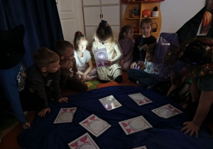 Dzieci siedzą w kole, w środku leżą obrazki. Dziewczynka trzyma obrazek i latarkę podczas wróżby z zawodami.