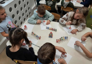 Dzieci siedzą przy stoliku i malują ceramiczne skarbonki w kształcie Mikołaja.