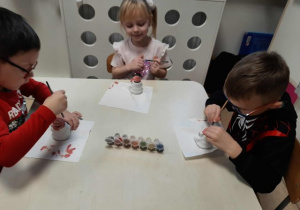 Dzieci siedzą przy stoliku i malują ceramiczne skarbonki w kształcie Mikołaja.