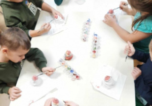 Przedszkolaki siedzą przy stoliku i malują ceramiczne skarbonki w kształcie Mikołaja.