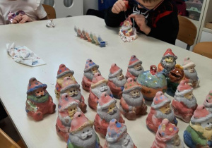 Chłopiec z dziewczynką siedzą przy stoliku i malują skarbonki. Na stole stoją pomalowane skarbonki.