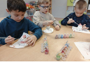 Dzieci siedzą przy stoliku i malują ceramiczne skarbonki Mikołaje