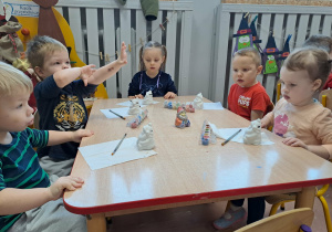 Dzieci siedzą przy stoliku i malują ceramiczne skarbonki Mikołaje