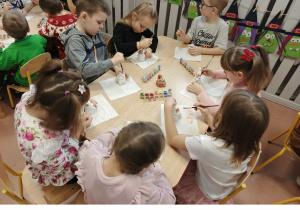 Dzieci siedzą przy stoliku i malują ceramiczne skarbonki Mikołaje