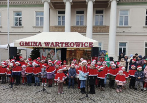 Przedszkolaki ubrane w czerwone peleryny i czapki śpiewają świąteczne piosenki.