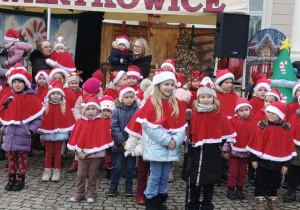Przedszkolaki ubrane w czerwone peleryny i czapki śpiewają świąteczne piosenki.