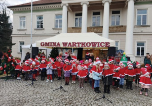 Przedszkolaki ubrane w czerwone peleryny i czapki śpiewają świąteczne piosenki.