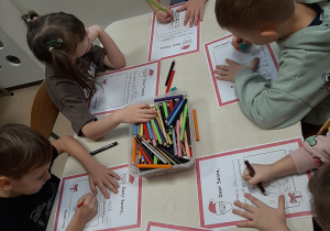 Kolejna grupa dzieci siedzi przy stoliku w sali przedszkolnej, rysują kredkami na kartce wymarzone prezenty.