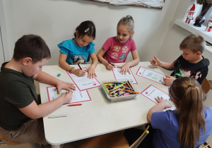 Następna grupa dzieci siedzi przy stoliku w sali przedszkolnej, rysują kredkami na kartce wymarzone prezenty.