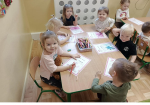 Kolejna grupa 3, 4 latków siedzi przy stoliku w sali przedszkolnej, rysują kredkami na kartce wymarzone prezenty.