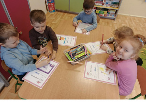 Następna grupa 3, 4 latków siedzi przy stoliku w sali przedszkolnej, rysują kredkami na kartce wymarzone prezenty.