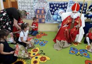 Dzieci rozpakowują prezenty od Mikołaja. W tle siedzi Mikołaj w stroju biskupa z Miry.