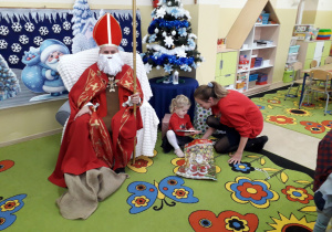 Mikołaj w stroju biskupa z Miry siedzi na fotelu i pozuje do zdjęcia. Obok dziewczynka z mamą rozpakowują prezent.