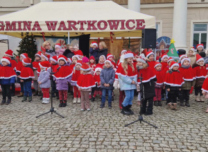 Dzieci ubrane w czerwone peleryny i czapki świąteczne śpiewają piosenki.