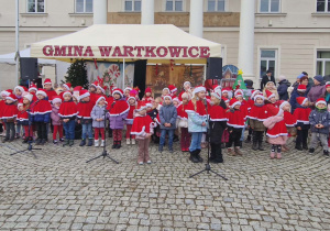 Dzieci ubrane w czerwone peleryny i czapki świąteczne śpiewają piosenki.