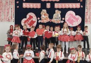 Przedszkolaki stoją na scenie, trzymają serca i recytują wierszyki. W tle dekoracja z okazji Dnia Babci i Dziadka.