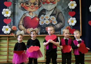 Dzieci recytują wiersz trzymając przed sobą czerwone serca z papieru.