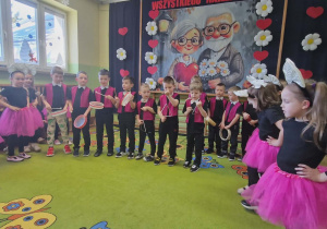 Dziewczynki stoją po bokach i śpiewają piosenkę. Chłopcy w środku grają na instrumentach.